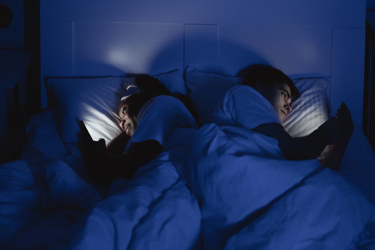 A couple lying back to back in bed at night, each looking at their own phone